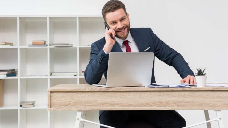 Hombre hablando por teléfono en una oficina sentado frente a un portatil situado en una mesa
